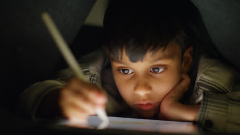 Close-Up-Of-Young-Boy-In-Home-Made-Camp-Made-From-Cushions-Playing-And-Drawing-With-Digital-Tablet-And-Stylus-At-Night-3